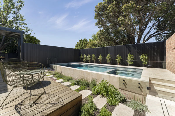 Elsternwick concrete pool and spa with travertine surrounds, pleached trees against a dark cladded fence and stepping stones through a herb garden by Fine Art Pools.